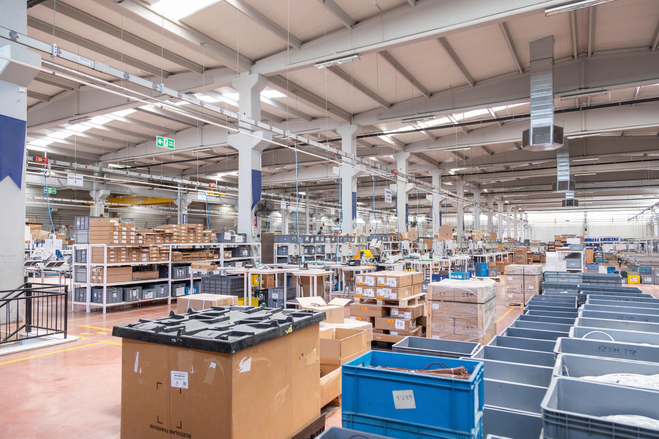 Spacious warehouse with shelves, stacked boxes, and bins under a high ceiling with industrial lighting.