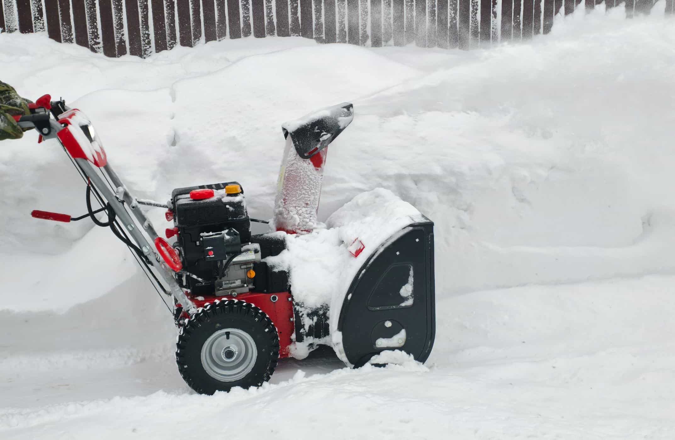 Person operating a red and black snowblower clearing deep snow near a wooden fence.
