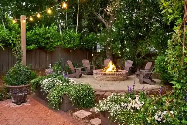 Cozy backyard patio with a lit fire pit surrounded by six Adirondack chairs, string lights overhead, and lush greenery and flowers in planters around the brick flooring.