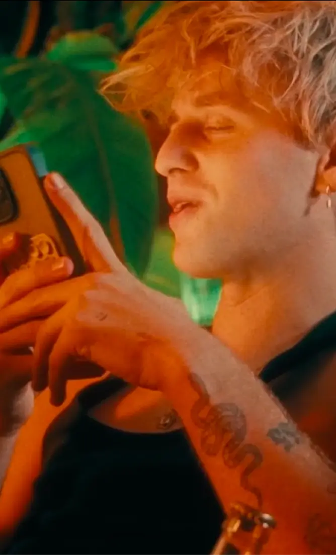 Young person with blonde hair and a snake tattoo on their arm looking at a phone in a warm-lit setting.