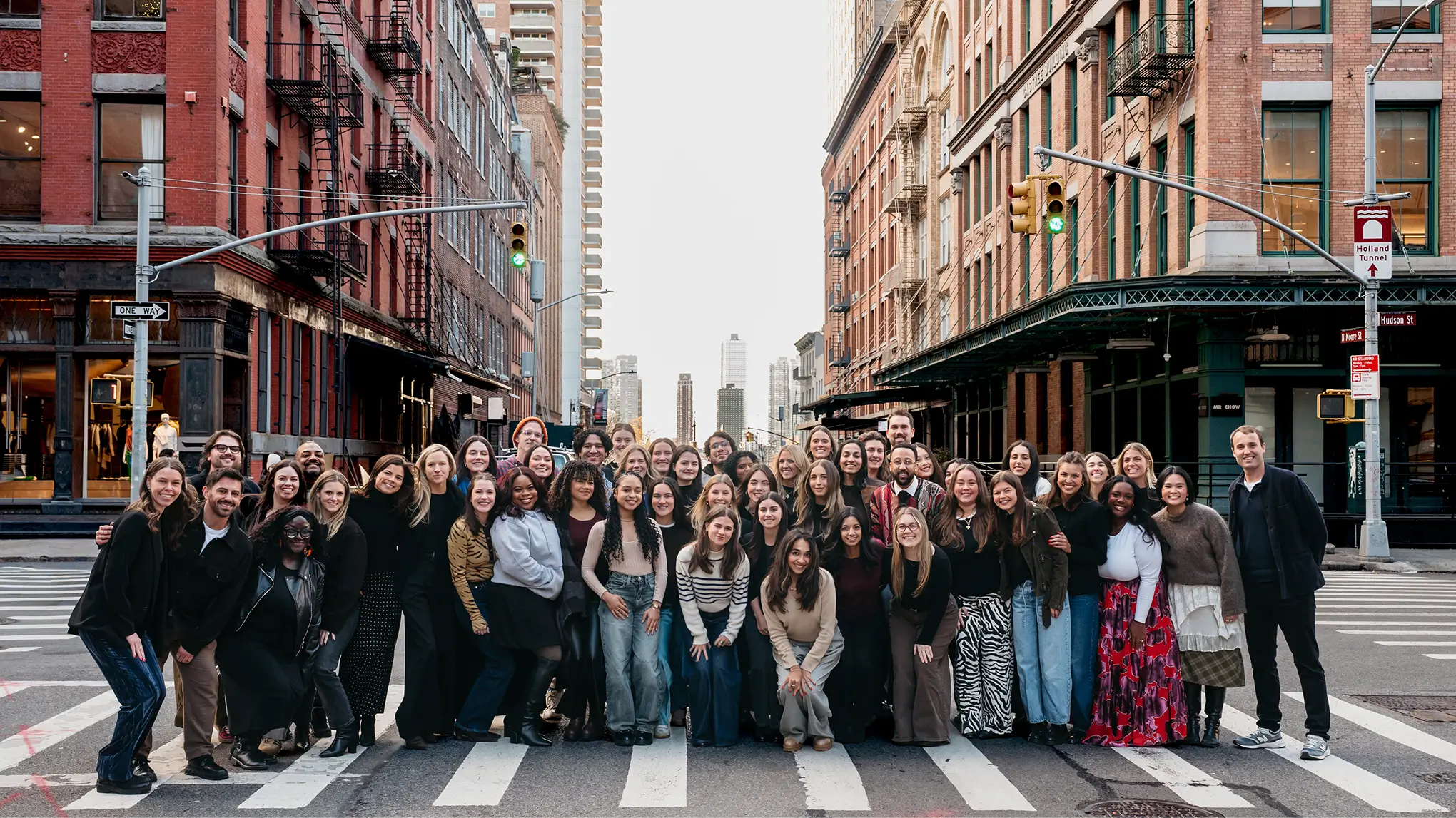 Large diverse group of adults posing smiling on a city street crosswalk with buildings and traffic lights in the background.