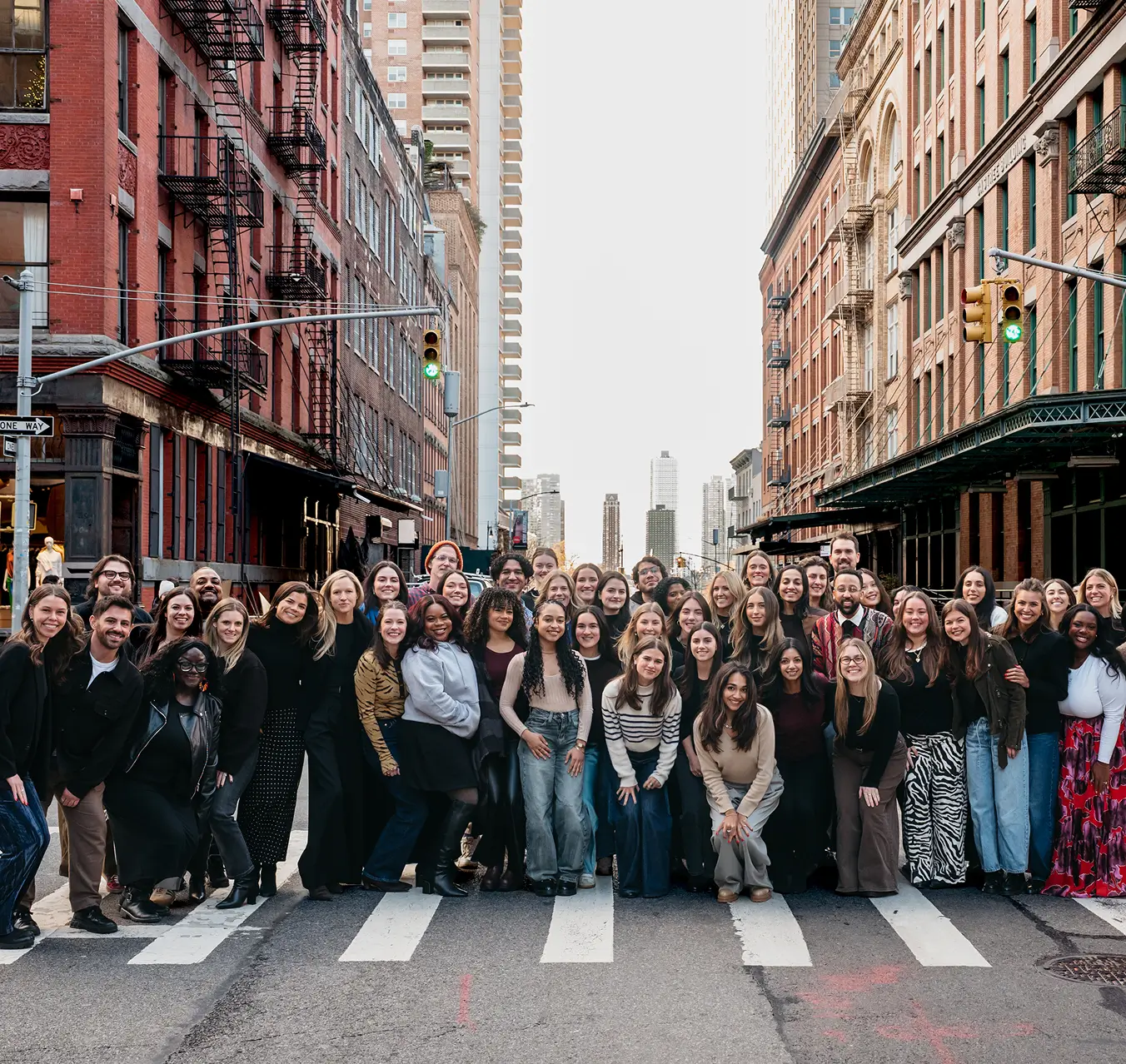 Large group of diverse people smiling and standing together on a crosswalk in a city street lined with brick buildings.