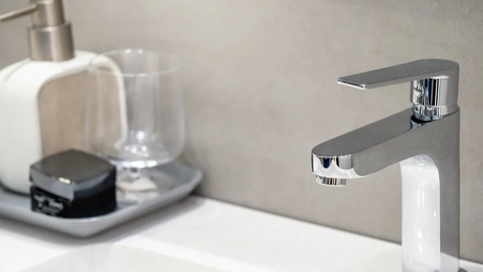 Modern chrome bathroom faucet on white sink with soap dispenser, tray, and clear glass in the background.