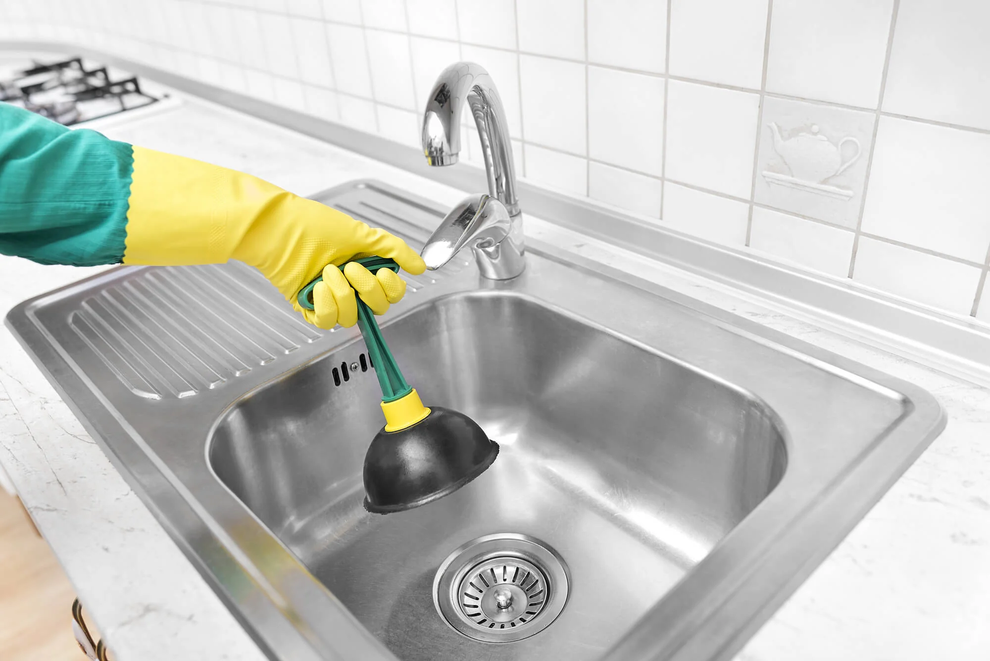 Person wearing yellow gloves using a plunger on a stainless steel kitchen sink.