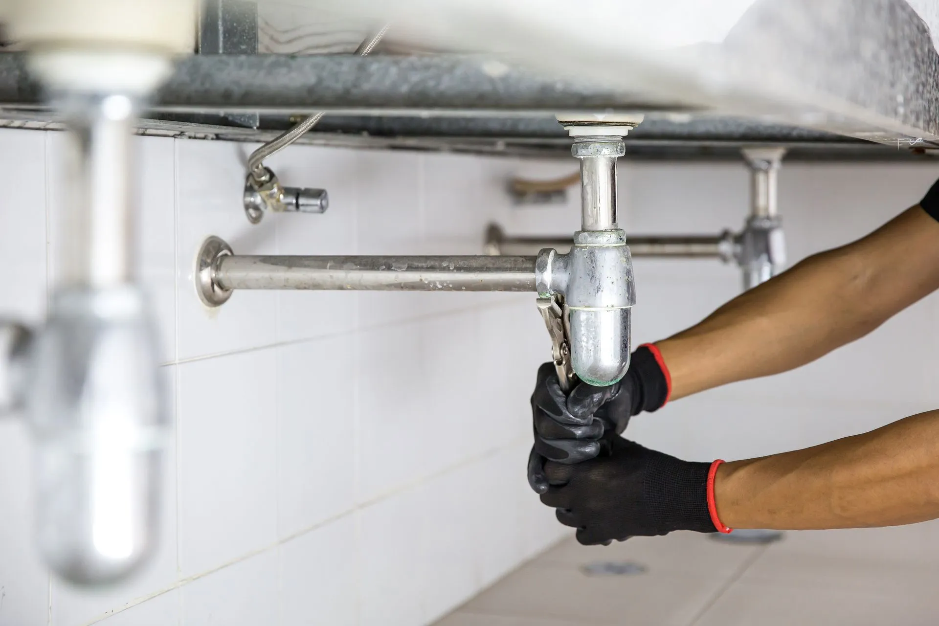 Person wearing black gloves using a wrench to tighten the pipe under a sink.