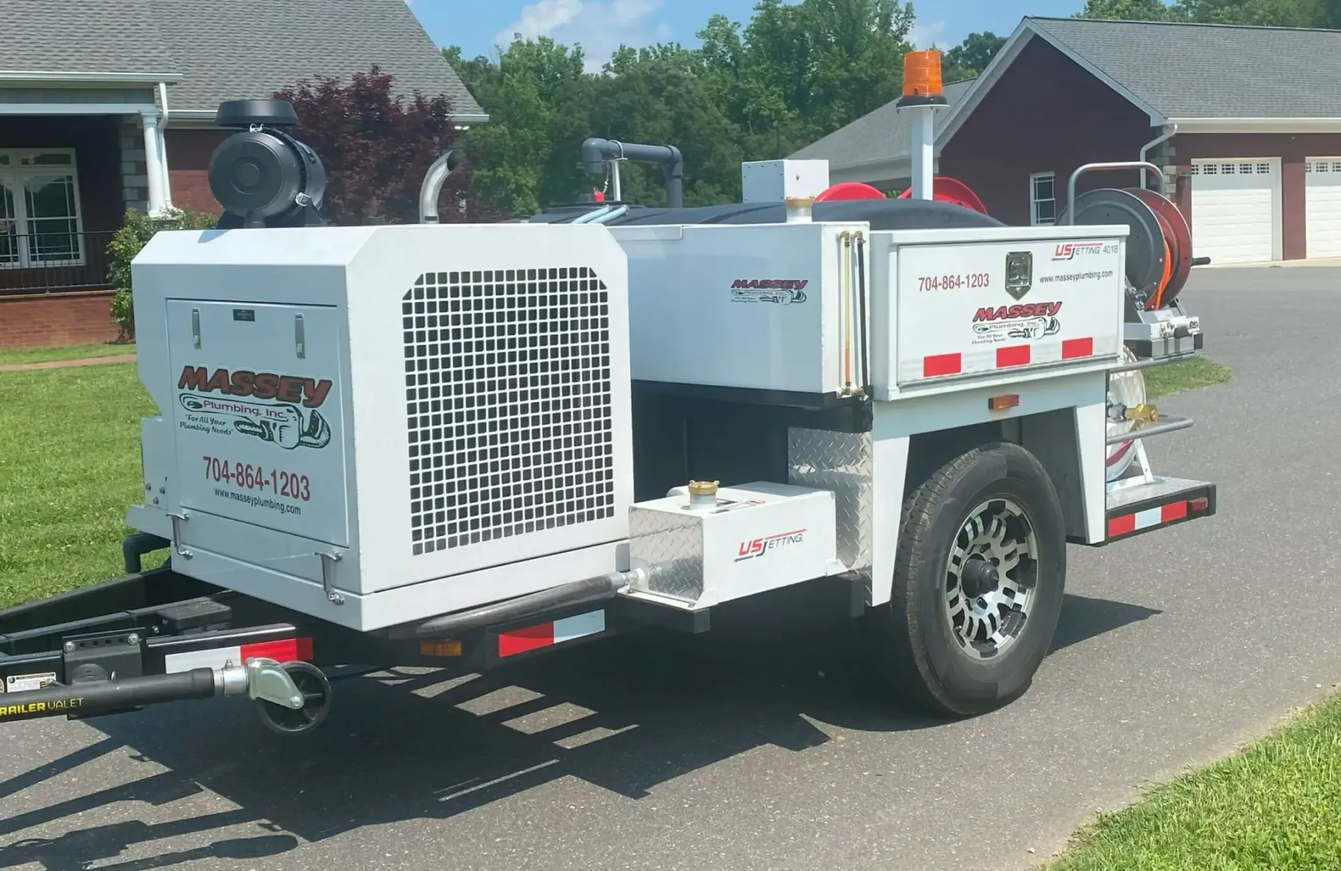White Massey Plumbing trailer-mounted pressure washer with contact number and website, parked on a driveway in a residential area.