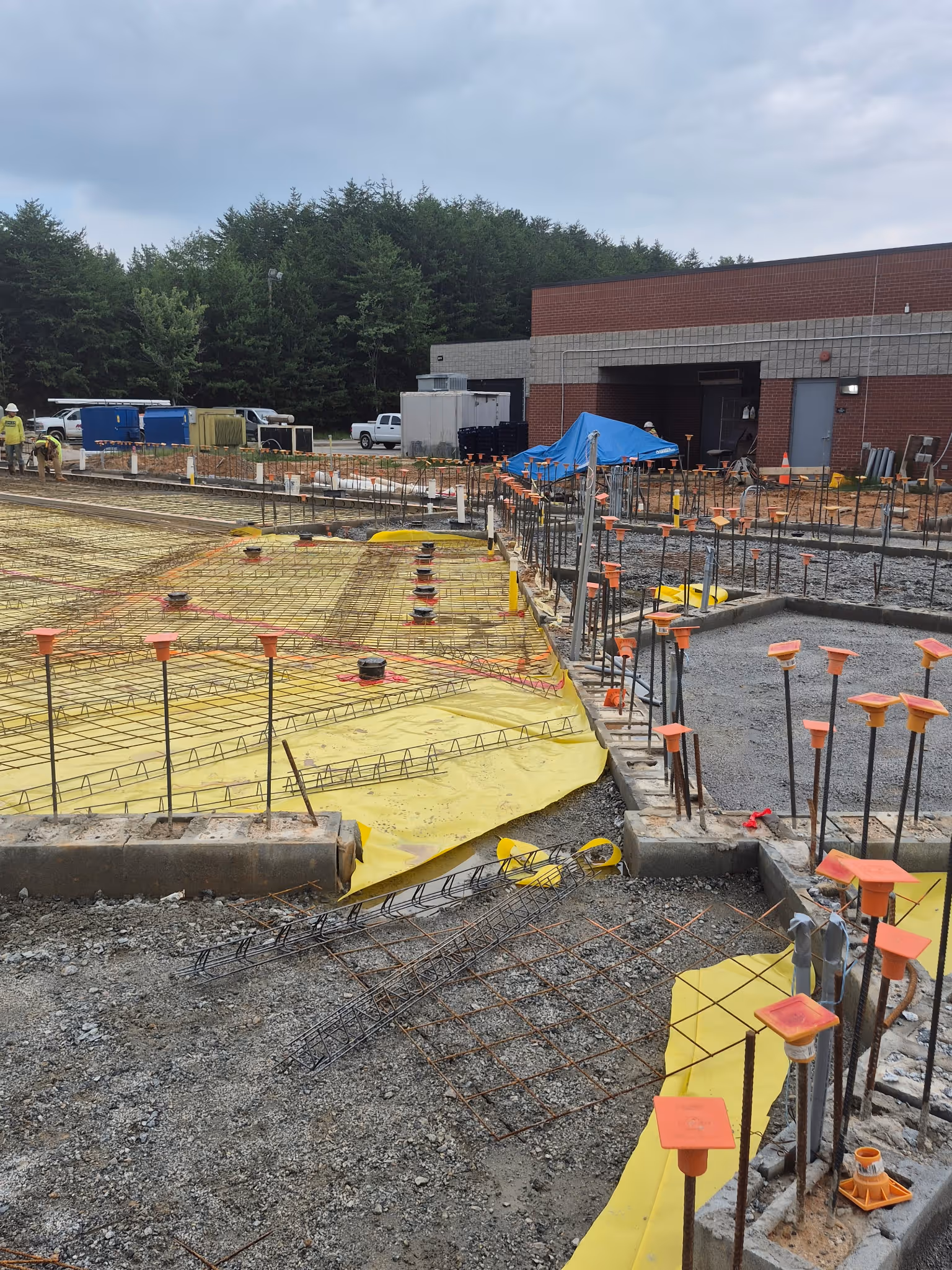 Construction site with foundation work in progress featuring steel rebar grids, yellow plastic sheeting, and safety caps on metal rods near a brick building.