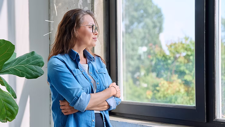 Woman in blue denim shirt gazes out a sunny window, arms crossed. Green leaves nearby. Calm, thoughtful atmosphere.