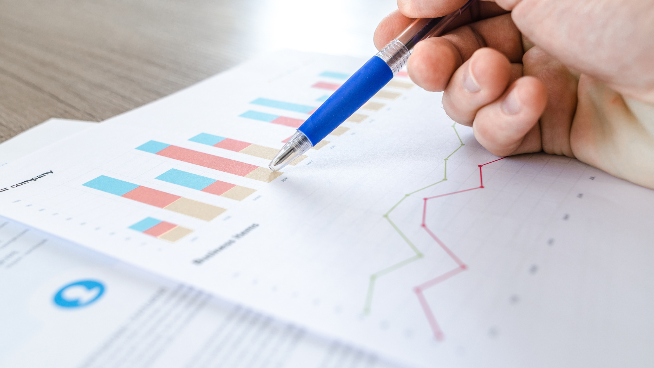 A hand holding a blue pen pointing at a bar chart and a line graph on a document.