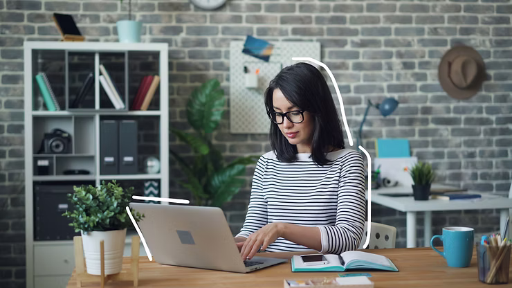 Woman in glasses types on a laptop at a wooden desk in a cozy home office with shelves, plants, and a brick wall.