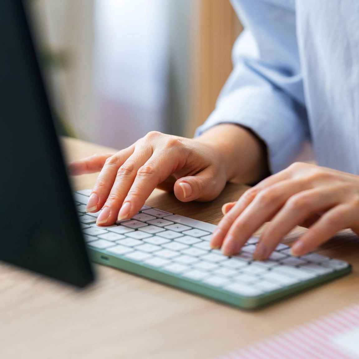 A close-up view of a person's hands typing on a wireless computer keyboard while sitting at a wooden desk