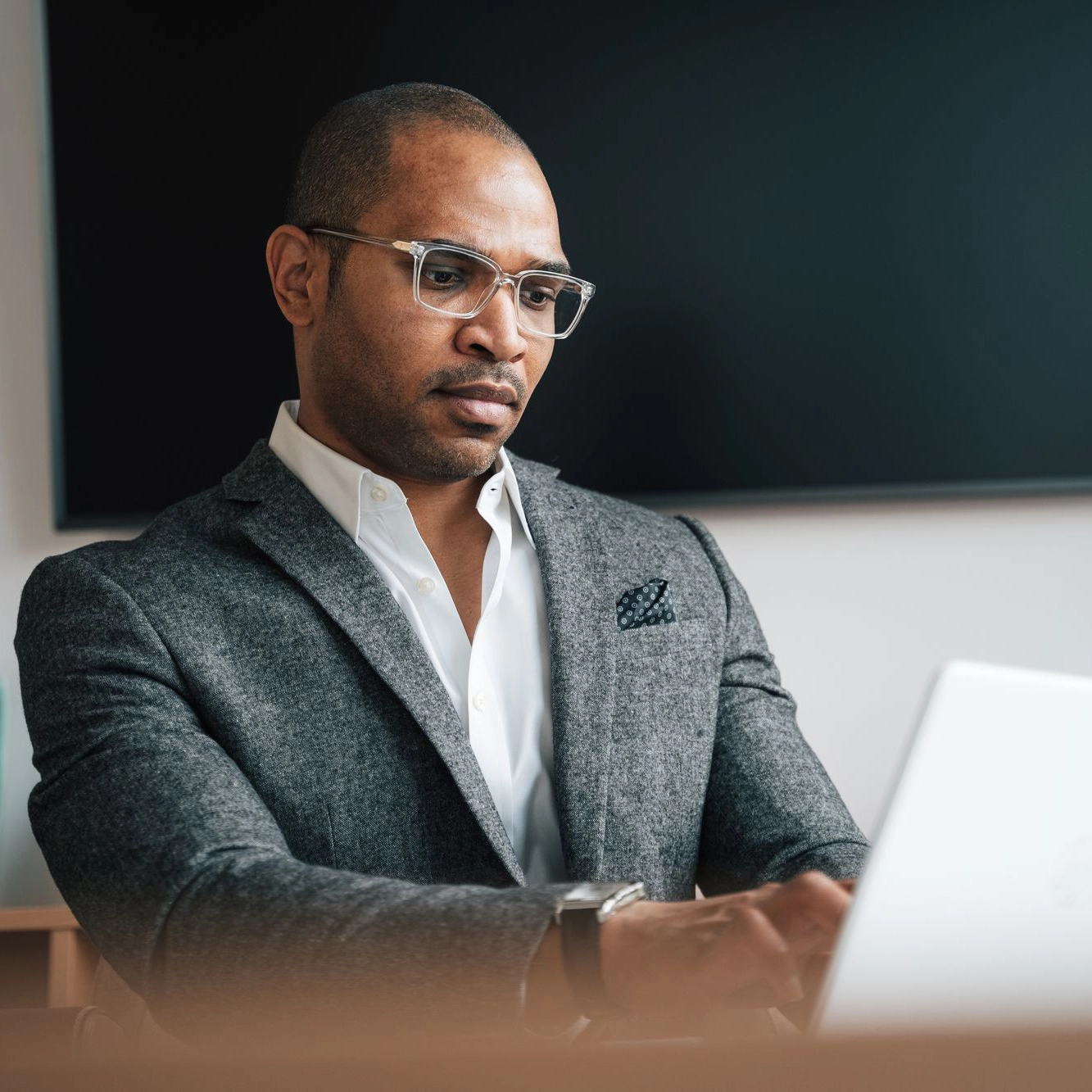 A professional man wearing clear-rimmed glasses and a gray tweed blazer sits in a corporate office focused on his laptop