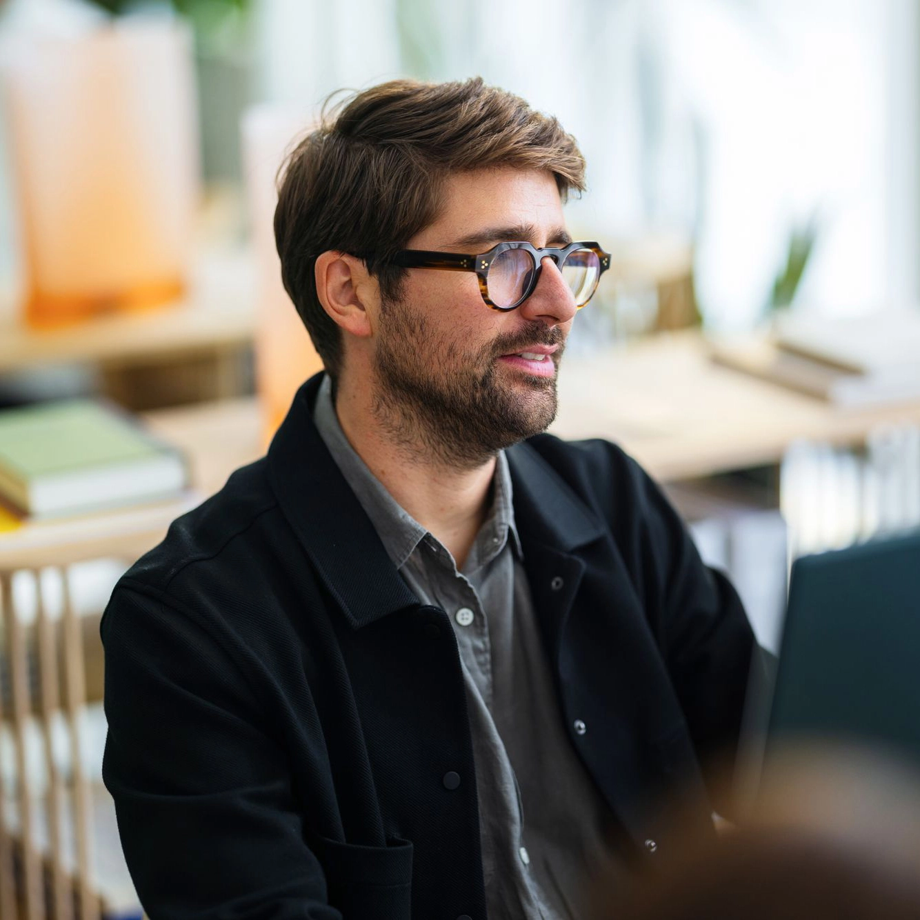 A bearded man wearing stylish glasses and a black jacket participates in a discussion in a modern office