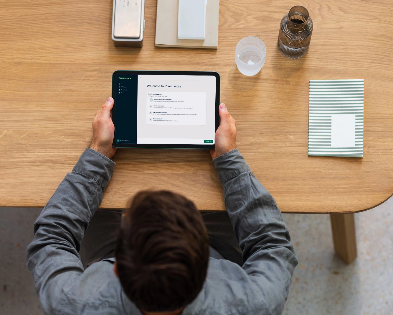 An overhead view of a man sitting at a wooden desk looking at the Promissory QSBS trust UI on a tablet computer
