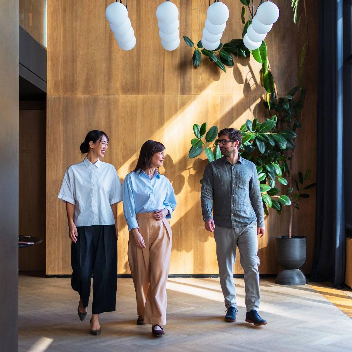 Three business colleagues walk and talk together through a modern, sunlit office with wood-paneled walls and stylish light fixtures
