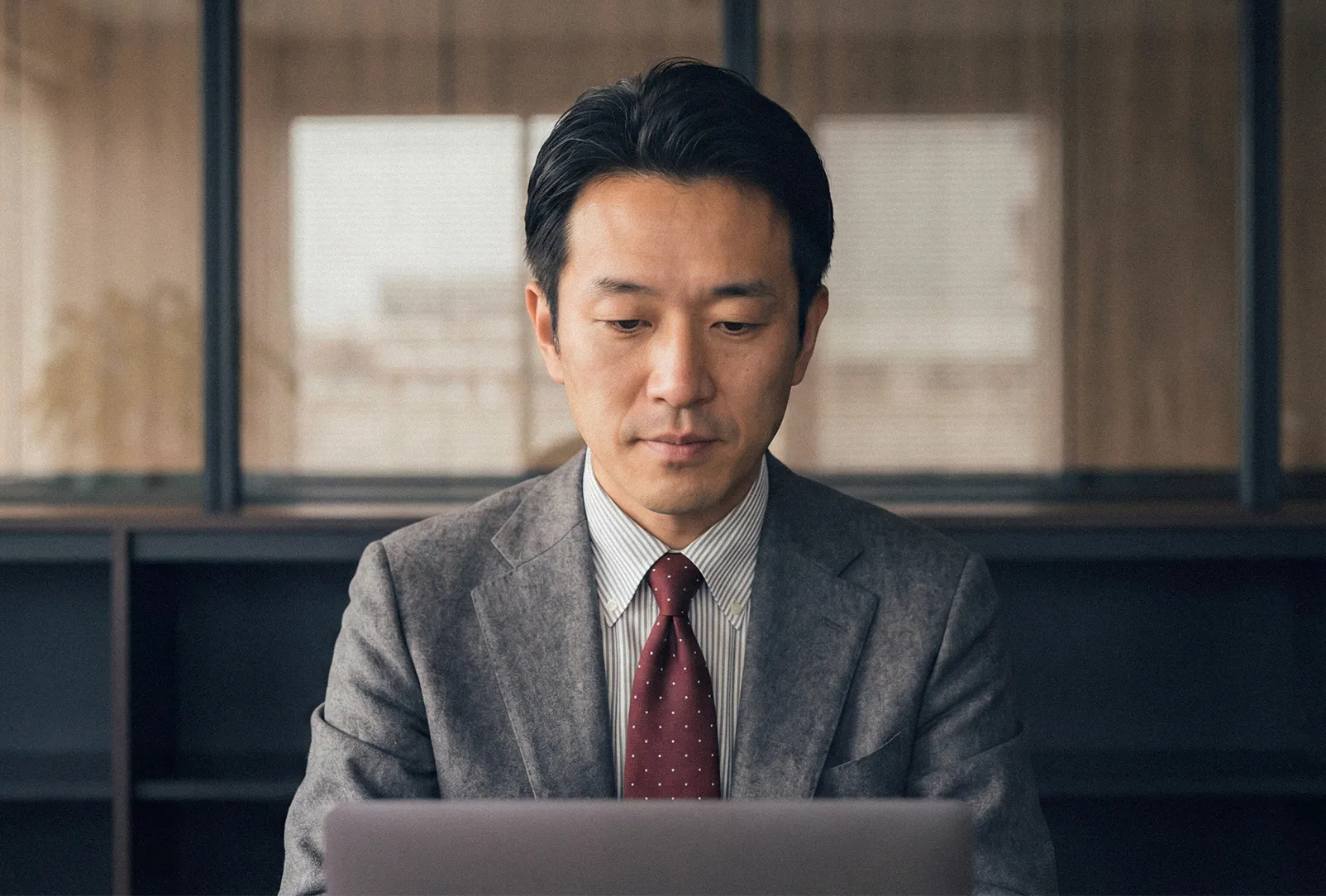 A professional man wearing clear-rimmed glasses and a gray tweed blazer sits in a corporate office focused on his laptop