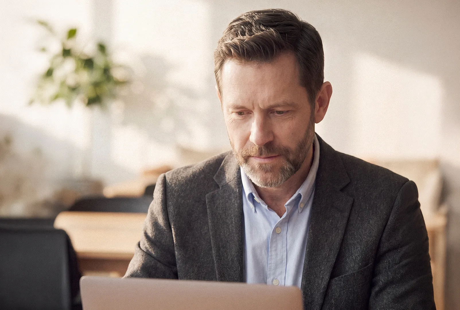 A professional man wearing clear-rimmed glasses and a gray tweed blazer sits in a corporate office focused on his laptop