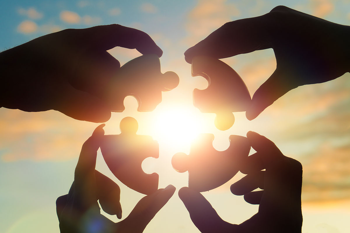Four hands putting together puzzle pieces in front of a bright sun