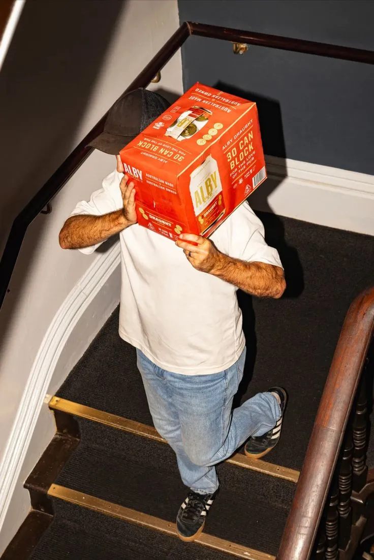 Person standing on carpeted stairs holding a red box of Alby Draught beer cans over their face.