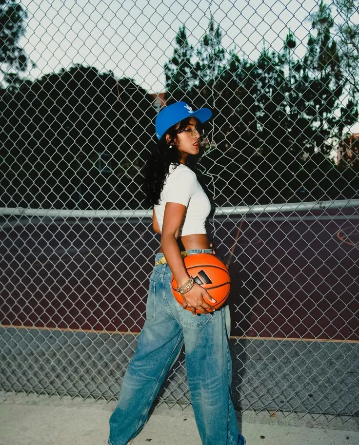 Young woman in a white crop top and blue cap holding a basketball, standing in front of a chain-link fence on a basketball court.