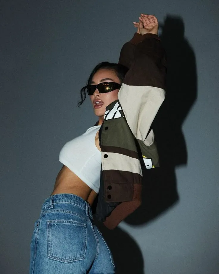 Woman wearing sunglasses, white crop top, blue jeans, and a color-blocked jacket, posing with arms raised against a gray background.