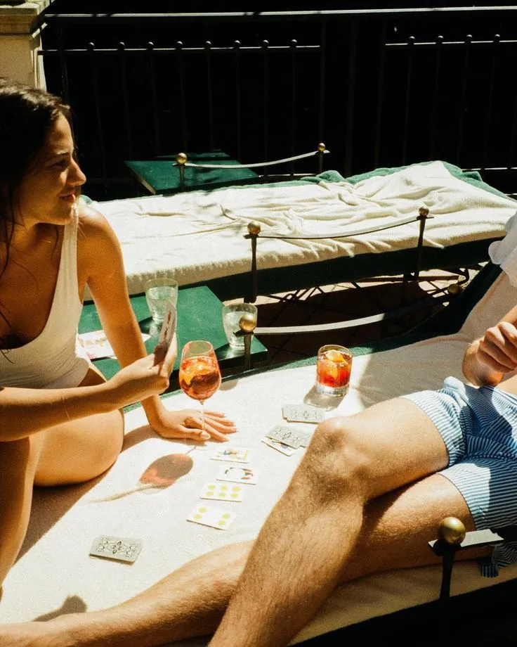 Two people playing cards and enjoying drinks on a sunbed outdoors in warm sunlight.