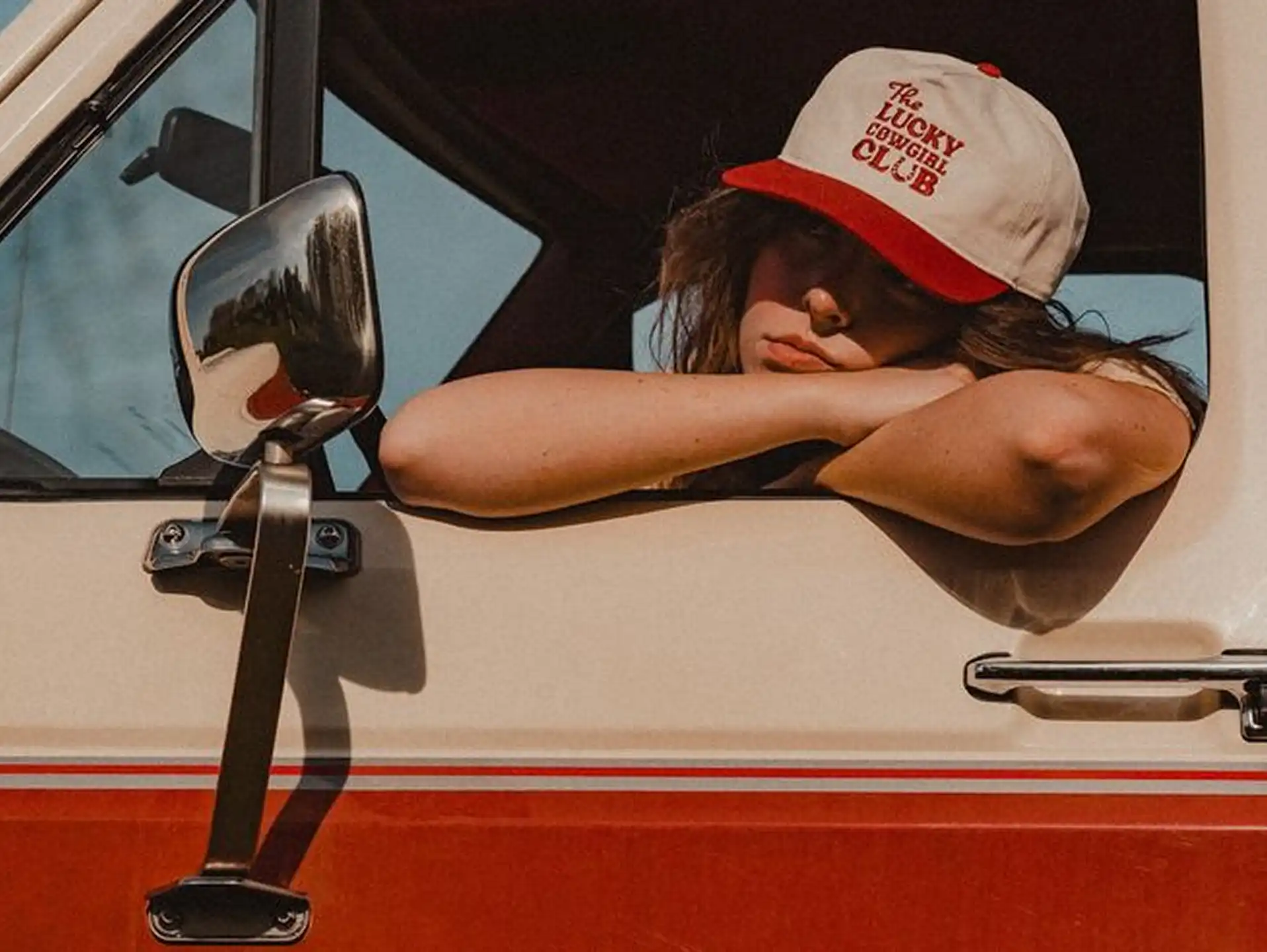 Person wearing a white and red cap with 'The Lucky Cowgirl Club' text leaning on the window of a two-tone red and white vehicle.