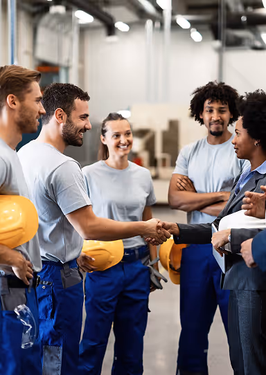 Factory workers in gray shirts and blue pants smiling while shaking hands with two professionals in a warehouse setting.
