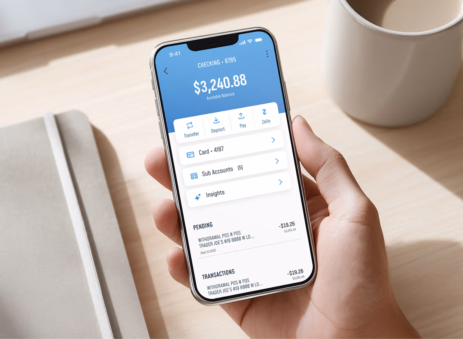 Hand holding smartphone showing banking app with checking account balance of $3,240.88 and recent transaction at Trader Joe's.
