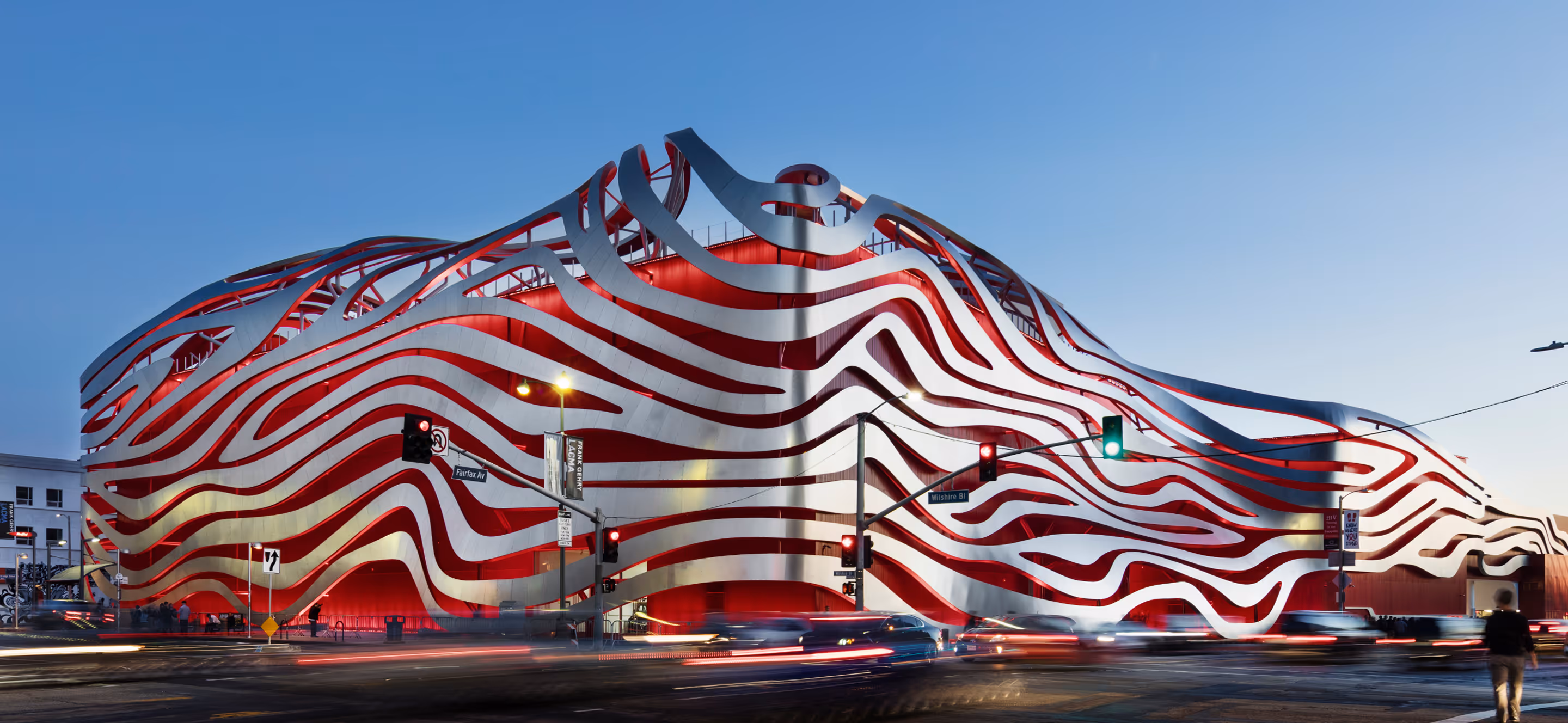 The Petersen Automotive Museum building with a red and white wavy metal facade illuminated at dusk at the intersection of Fairfax Ave and Wilshire Blvd.