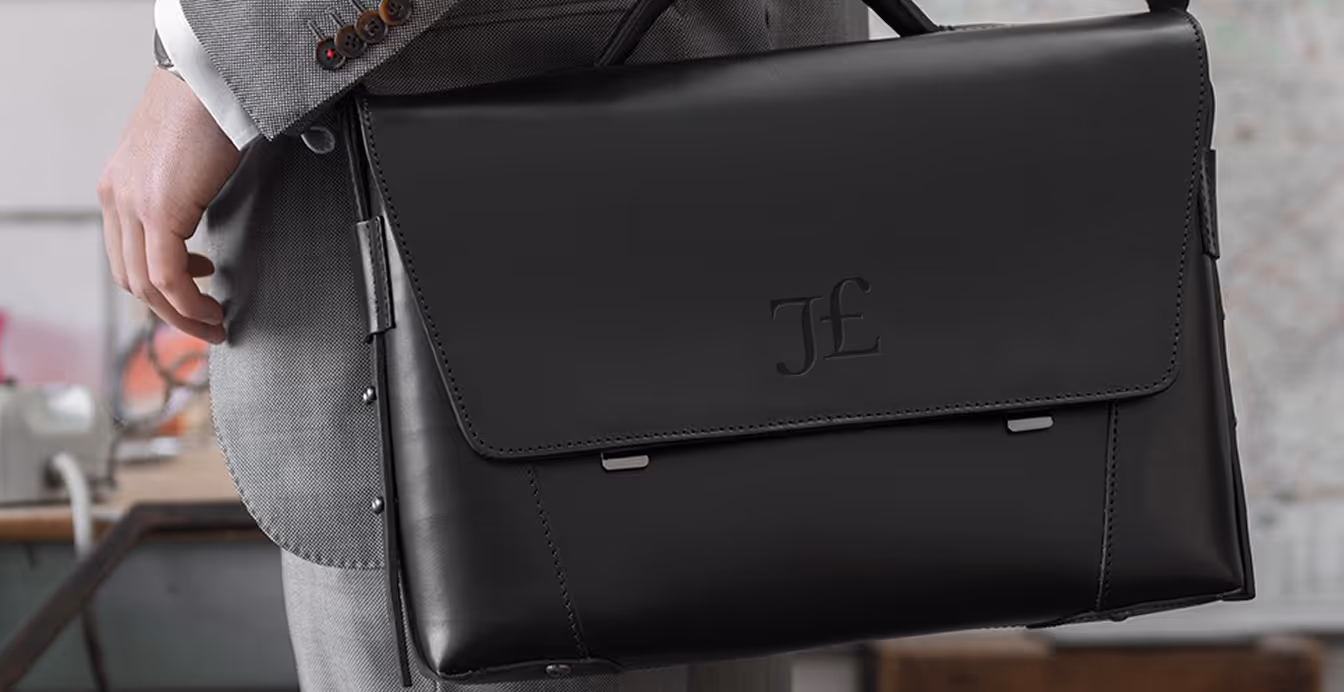 Person in gray suit holding a black leather briefcase with embossed JL initials on the flap.
