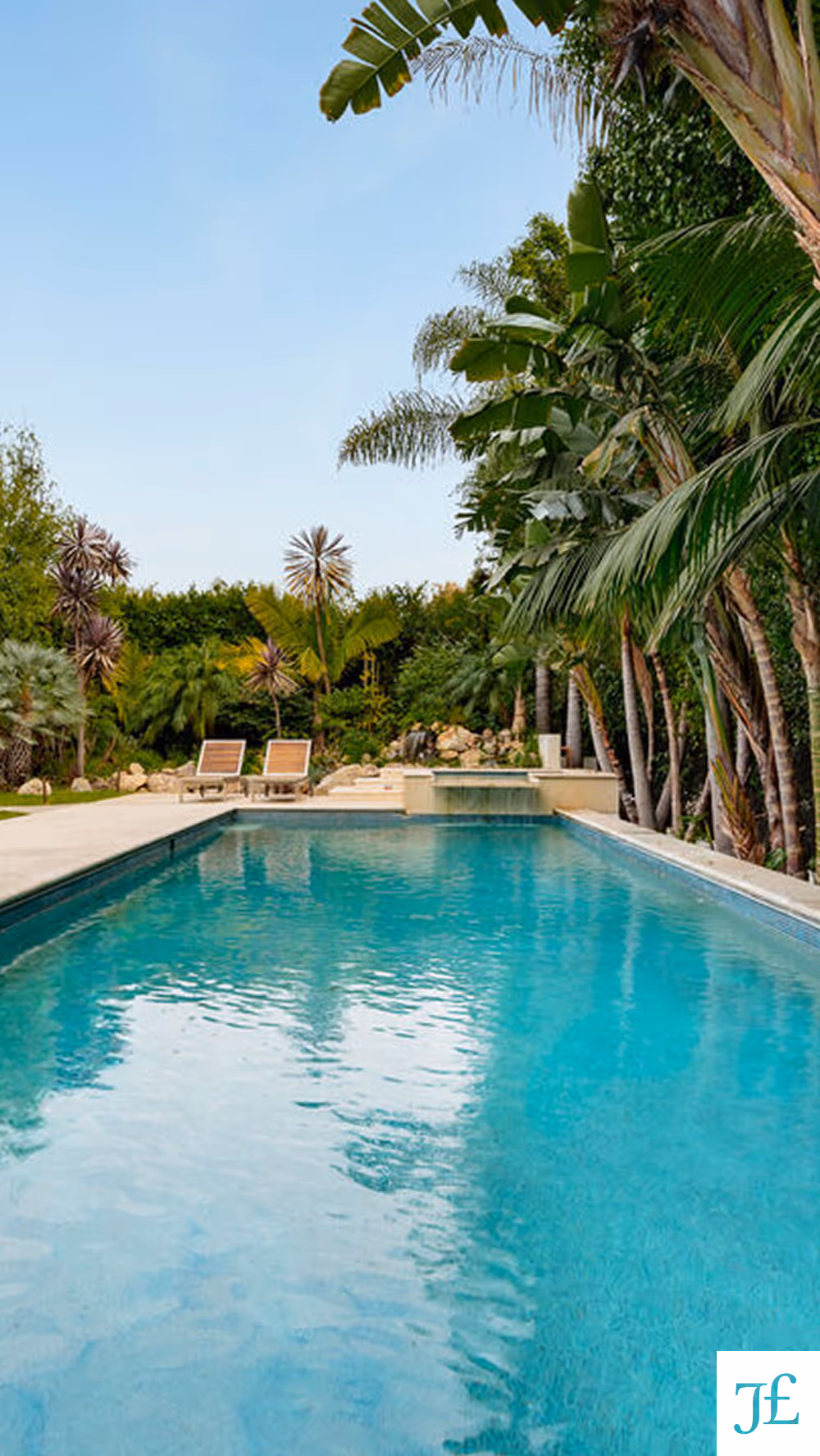 Clear blue swimming pool surrounded by tropical palm trees and two lounge chairs under a clear sky.