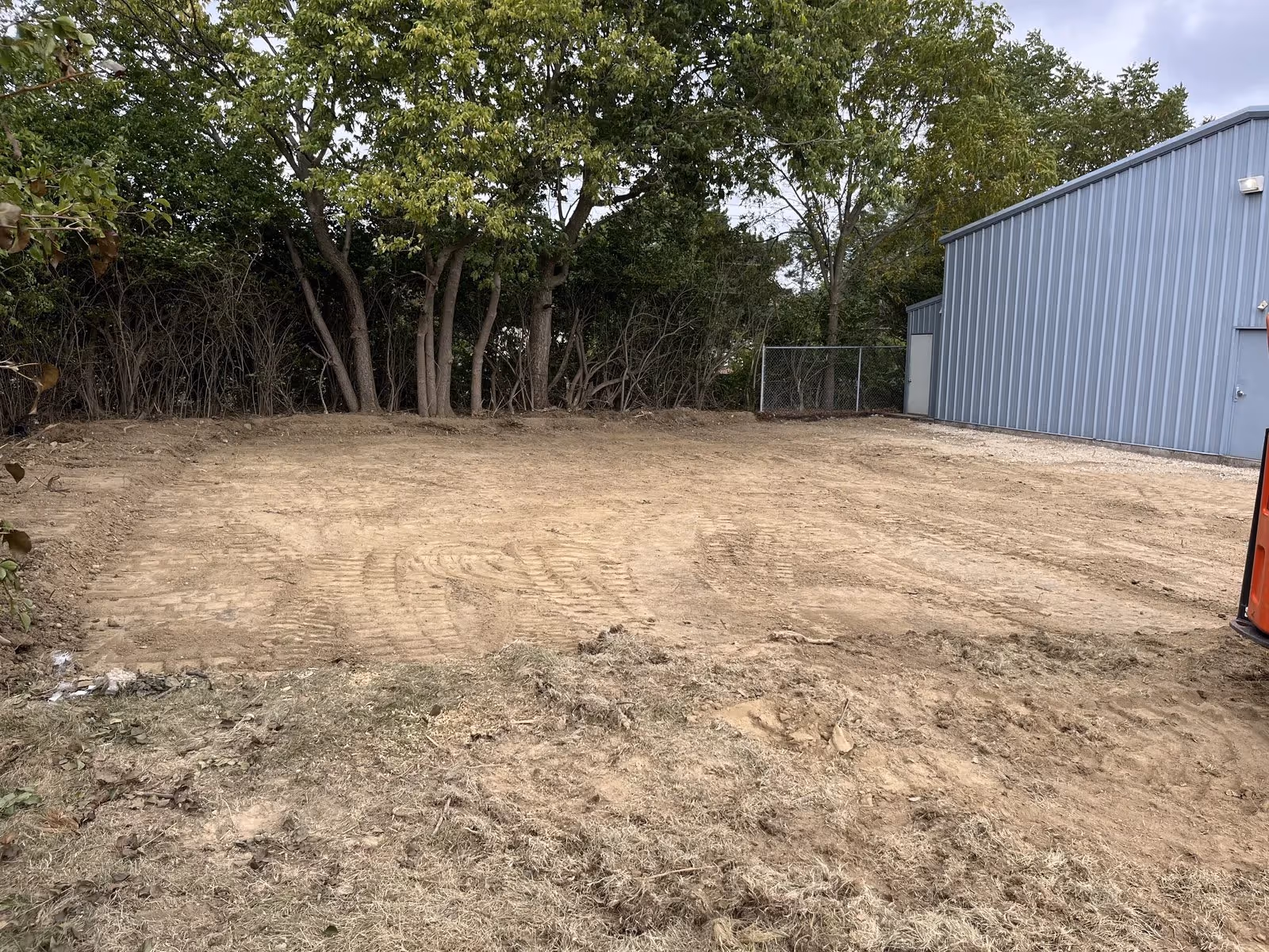 Cleared dirt lot with tire tracks next to a blue metal building and surrounded by trees under a cloudy sky.