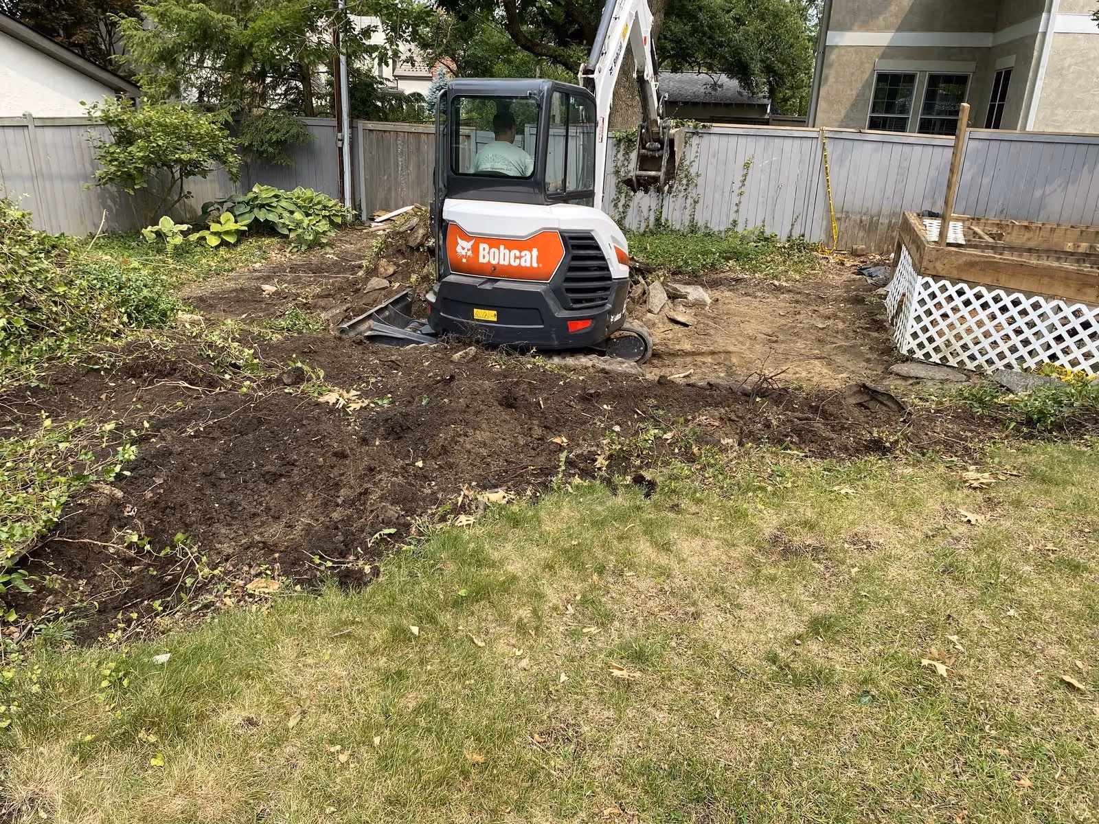 Bobcat excavator digging soil in a backyard garden near a wooden deck and fenced area.