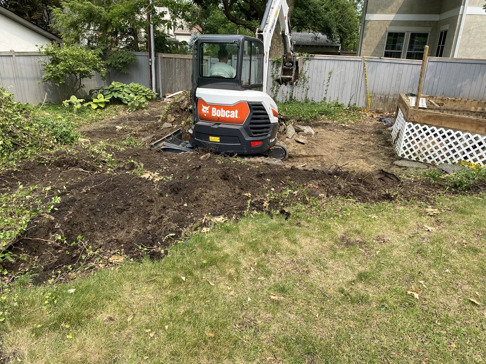 Bobcat excavator digging soil in a backyard garden near a wooden deck and fenced area.
