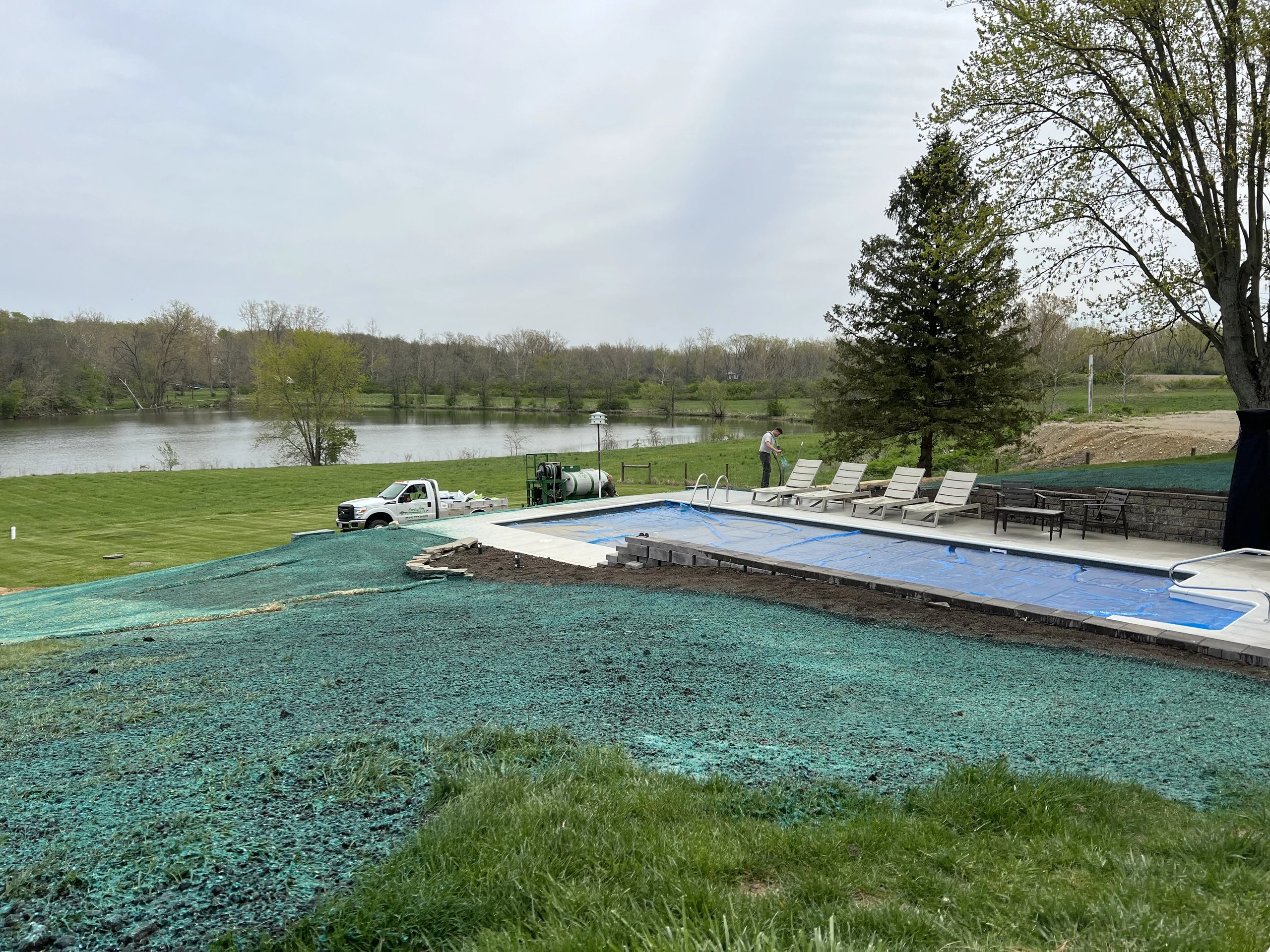 Outdoor pool covered with a blue tarp beside a patio with lounge chairs and a lake in the background.
