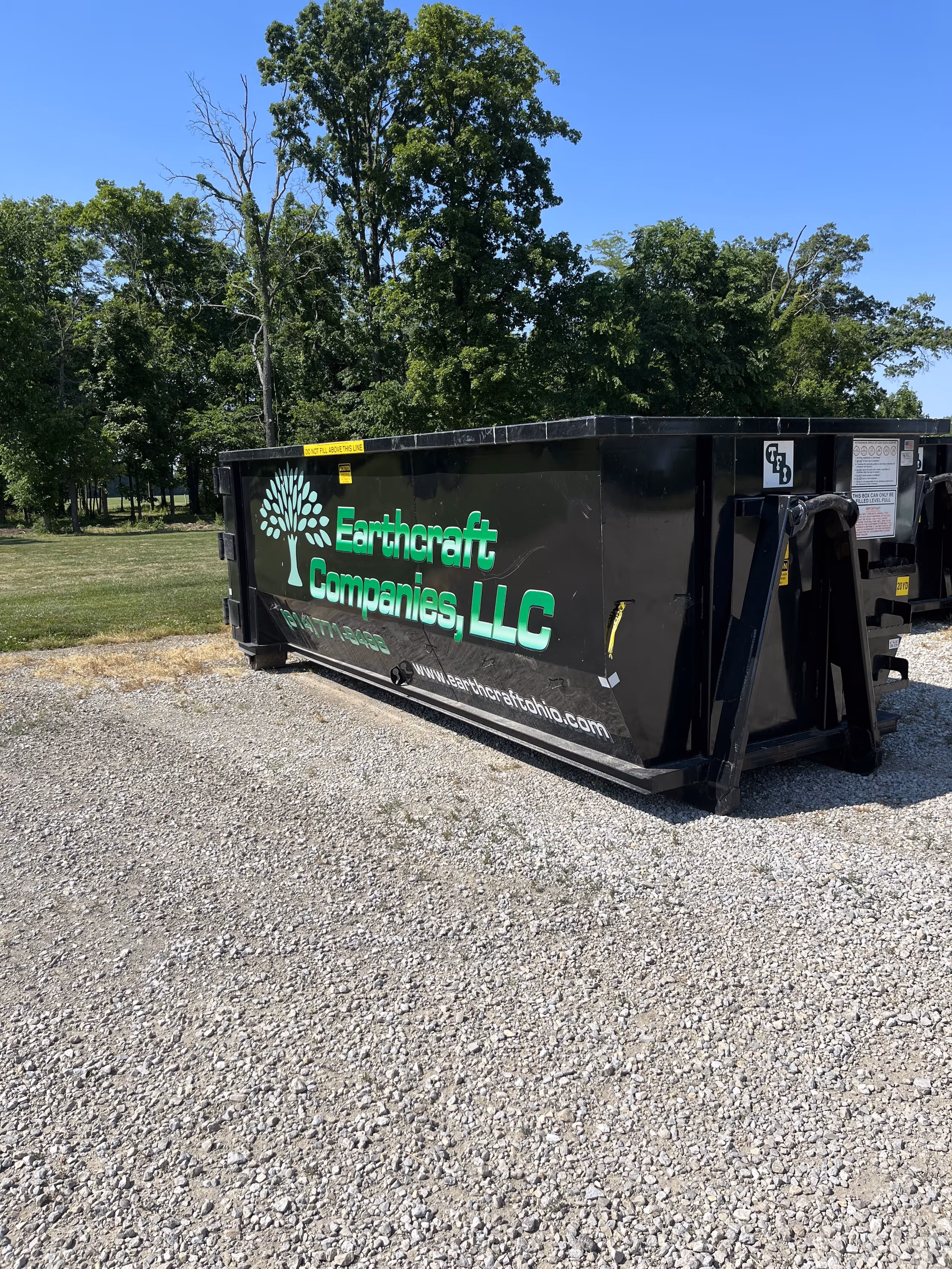 Black dumpster with green text and a tree logo for Earthcraft Companies, LLC, on gravel with trees and blue sky in the background.