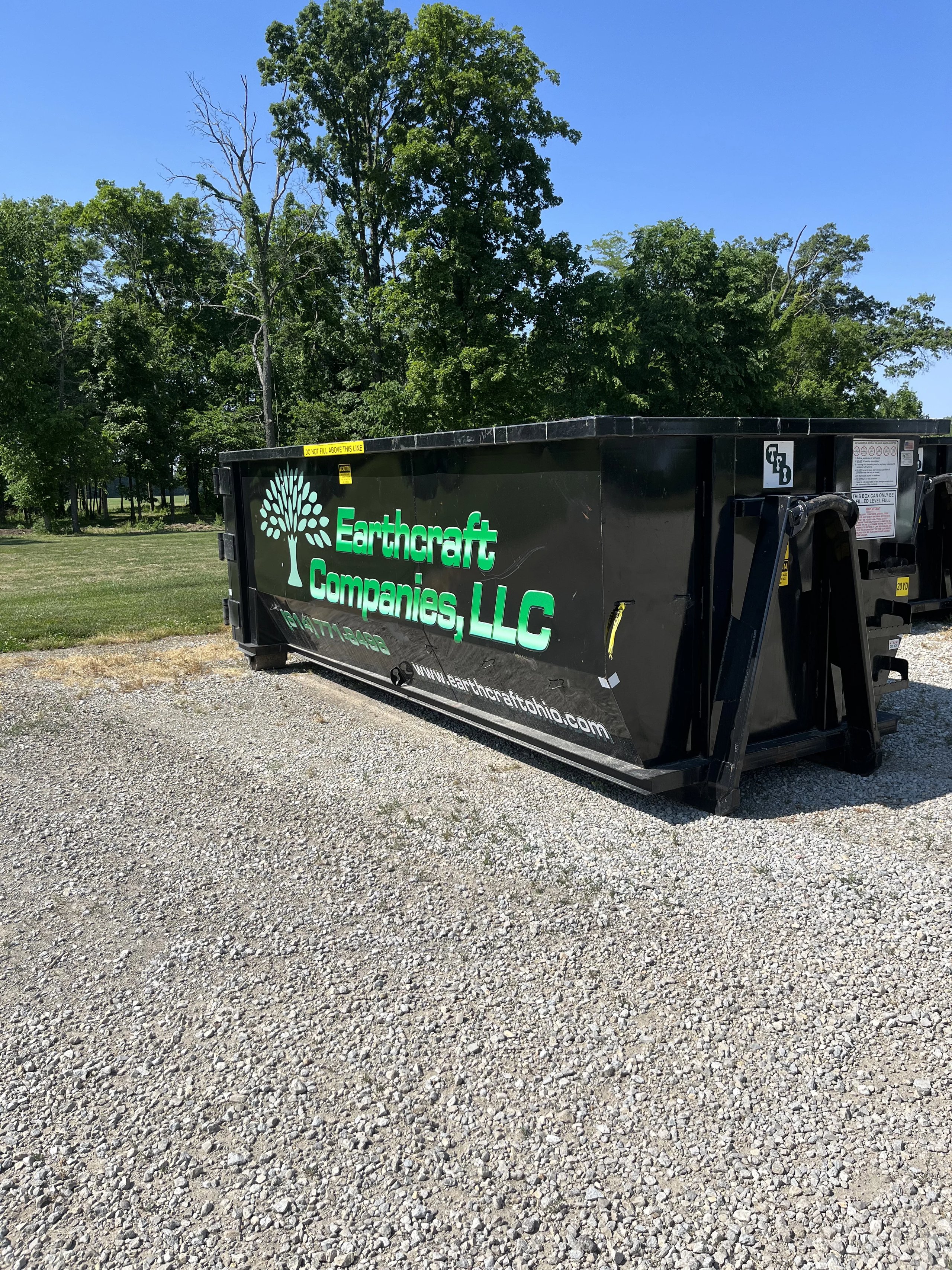 Black dumpster with green text and a tree logo for Earthcraft Companies, LLC, on gravel with trees and blue sky in the background.
