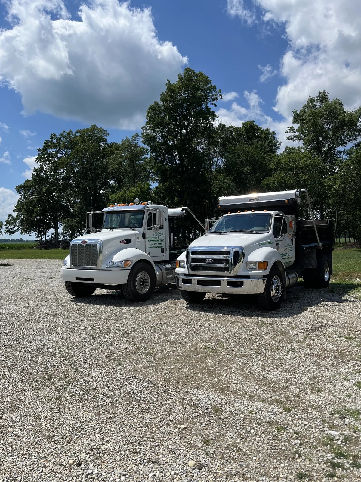 Two white commercial trucks parked on a gravel lot with trees and a partly cloudy sky in the background.