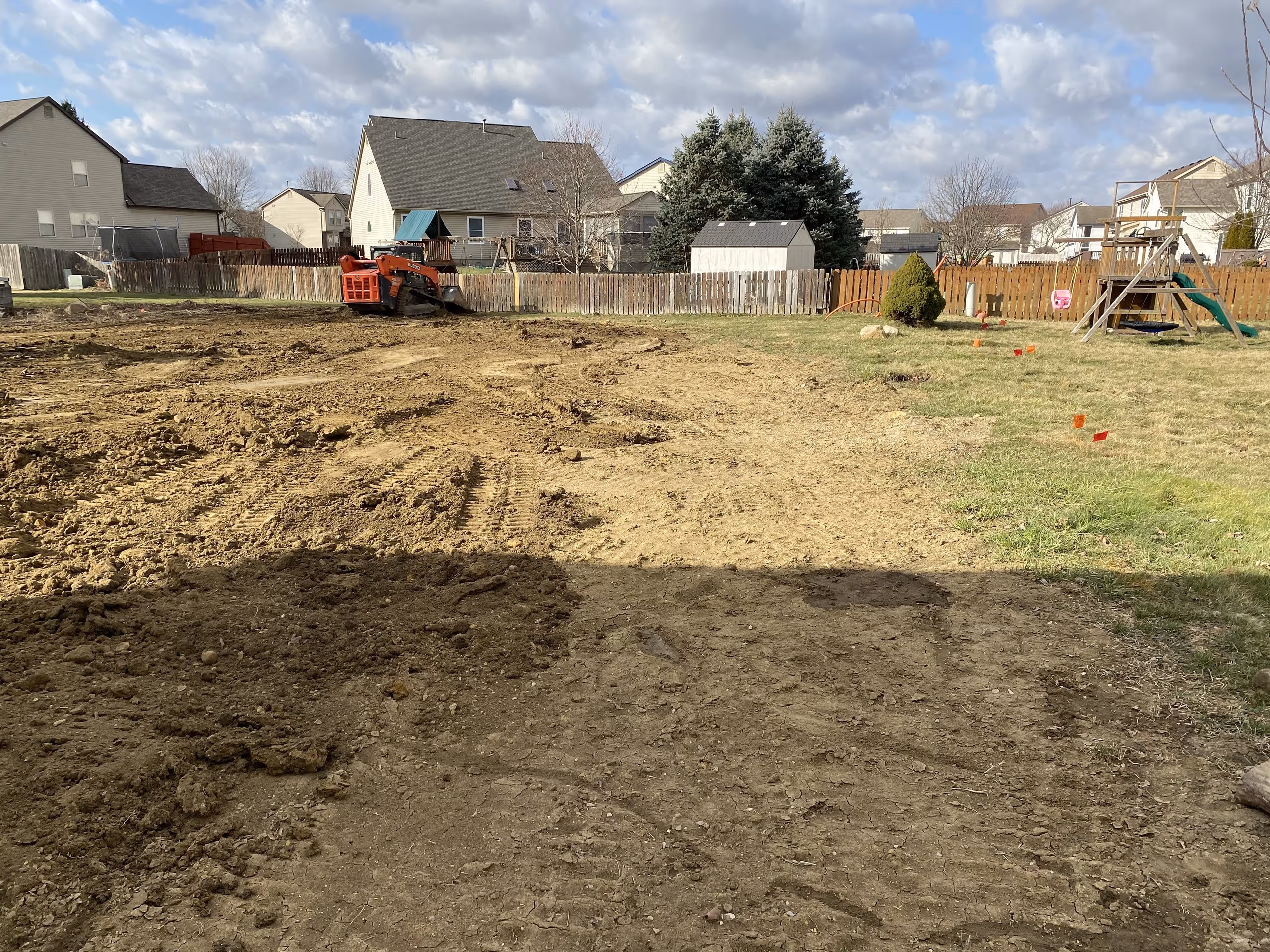 Residential backyard with a cleared and leveled dirt area and a small orange bulldozer near the wooden fence surrounded by houses.