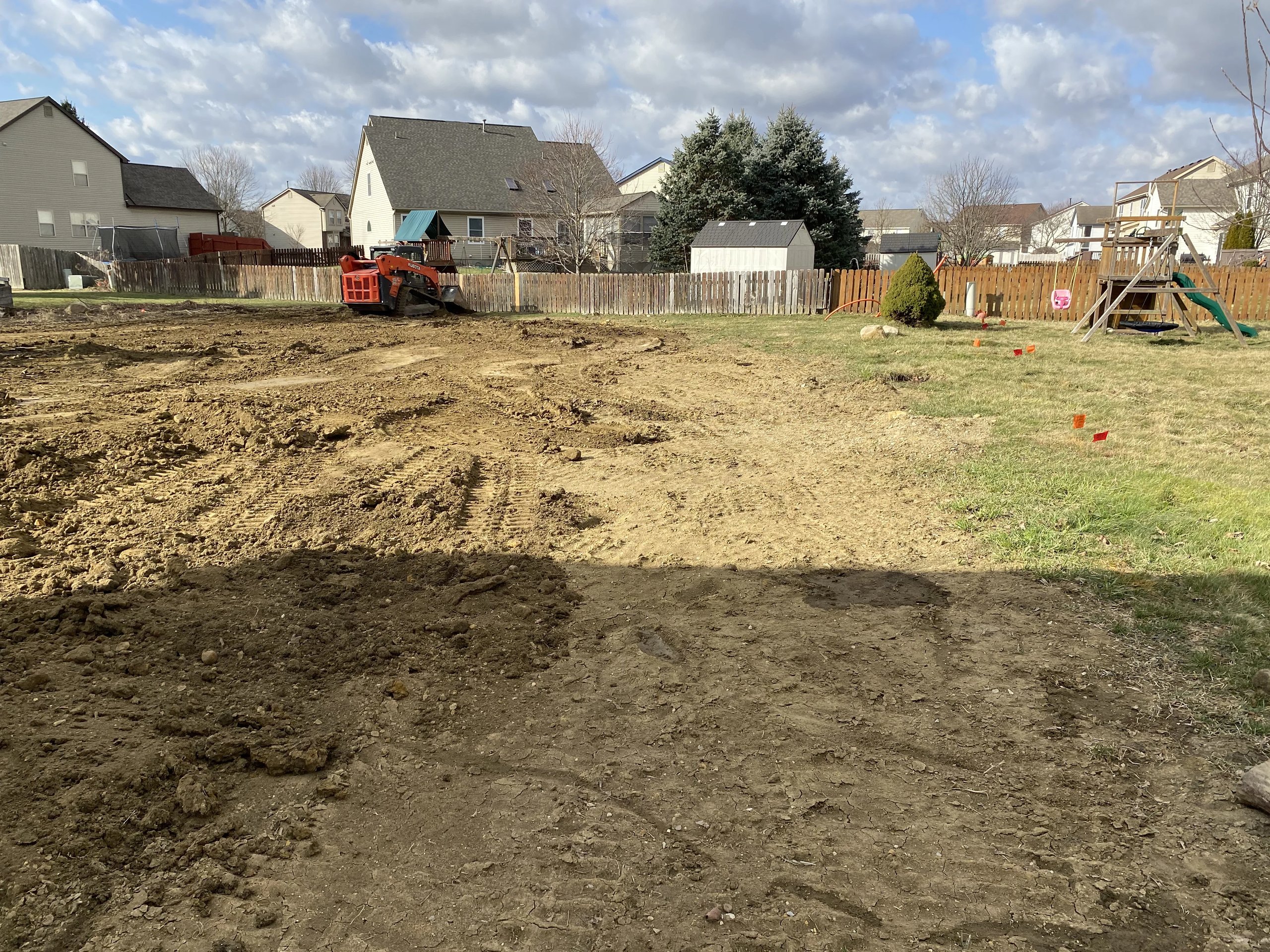 Residential backyard with a cleared and leveled dirt area and a small orange bulldozer near the wooden fence surrounded by houses.