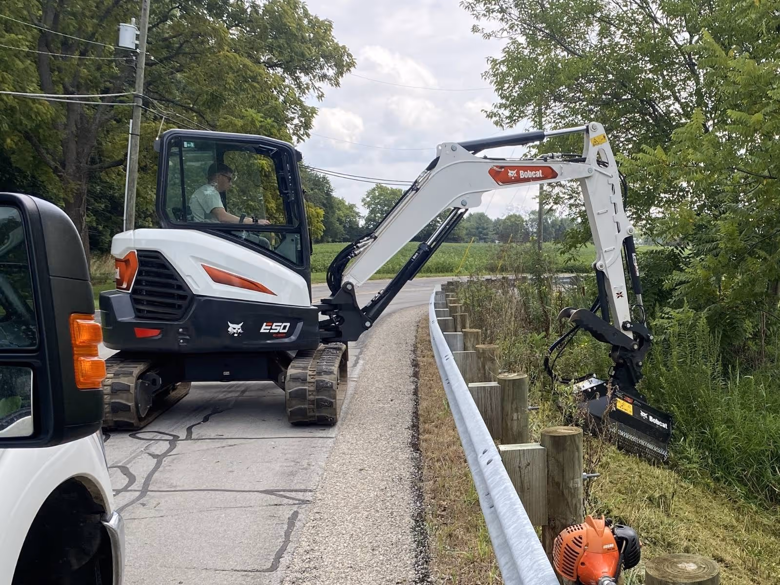 Bobcat E50 compact excavator clearing vegetation beside road guardrail with a flail mower attachment.