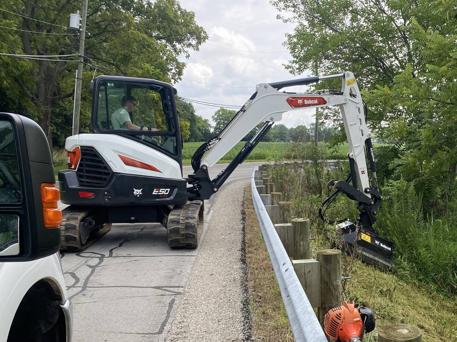 Bobcat E50 compact excavator clearing vegetation beside road guardrail with a flail mower attachment.