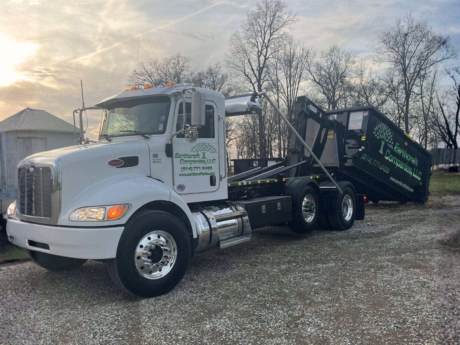 White roll-off truck lifting a large black dumpster container with Eartherott Companies LLC branding parked on gravel with leafless trees in the background.