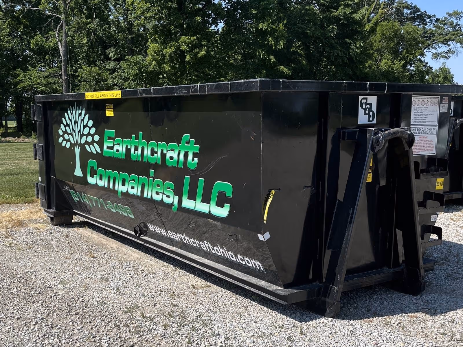 Black roll-off dumpster with green text reading 'Earthcraft Companies, LLC' and a tree logo, placed on gravel with trees in the background.