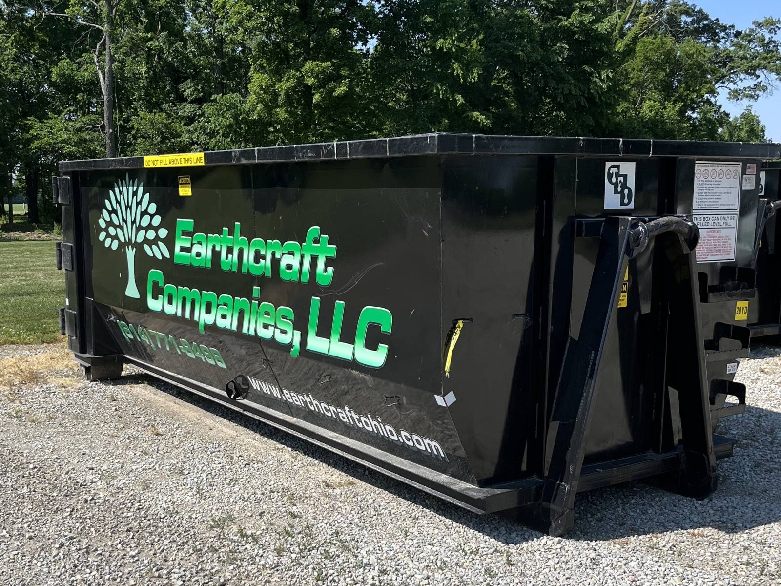 Black roll-off dumpster with green text reading 'Earthcraft Companies, LLC' and a tree logo, placed on gravel with trees in the background.