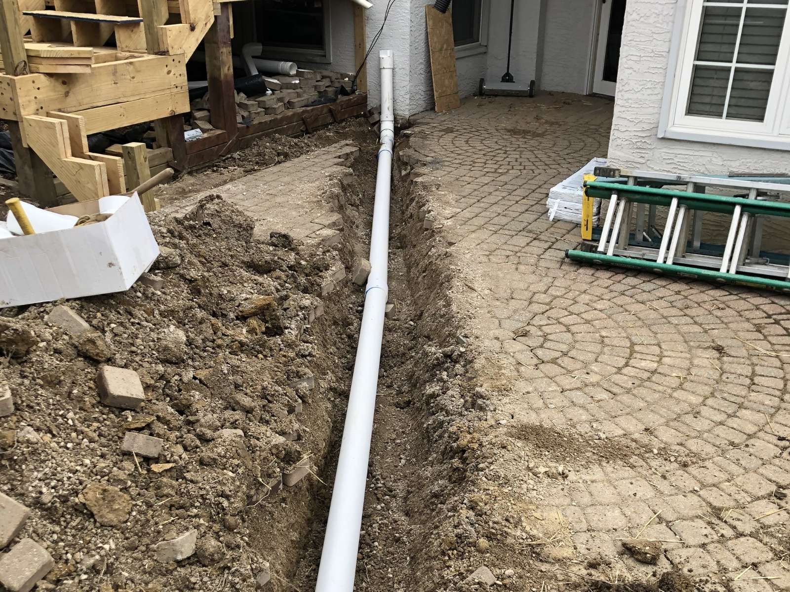 PVC drainage pipe laid in a trench beside a house with patterned brick patio and construction materials nearby.