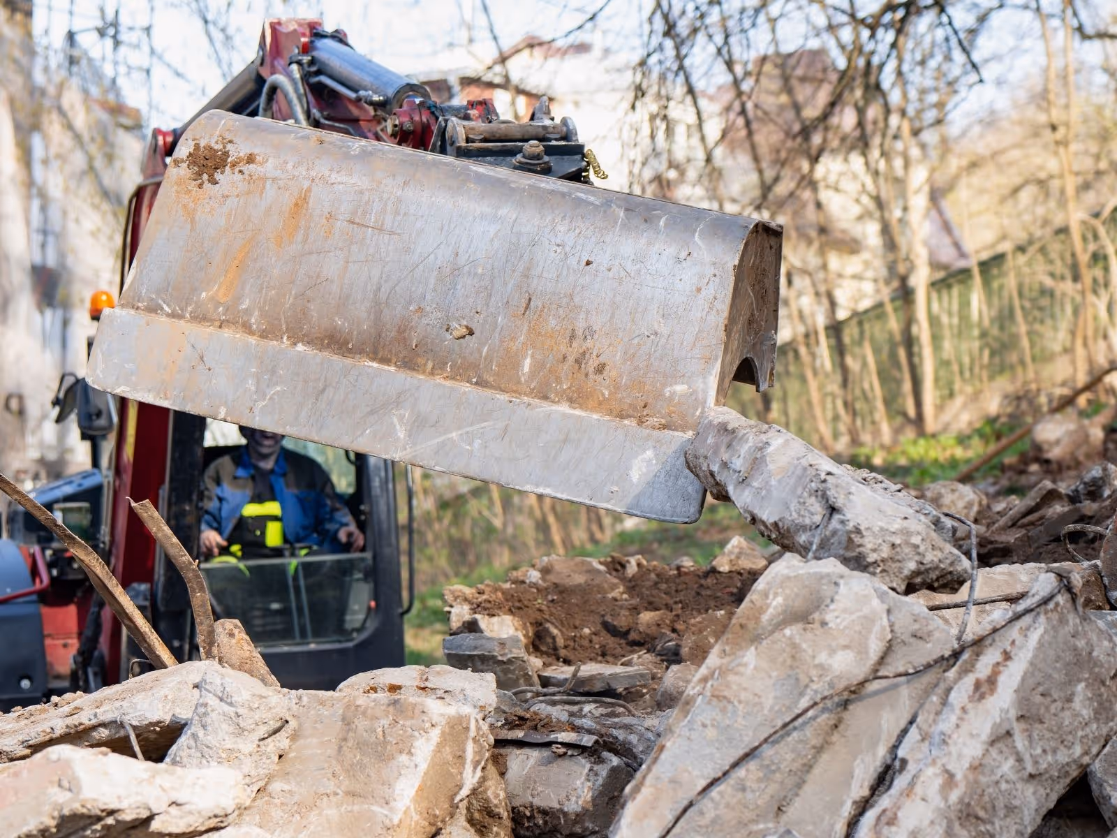 Excavator bucket lifting and moving broken concrete rubble at a demolition site with operator visible in the cabin.