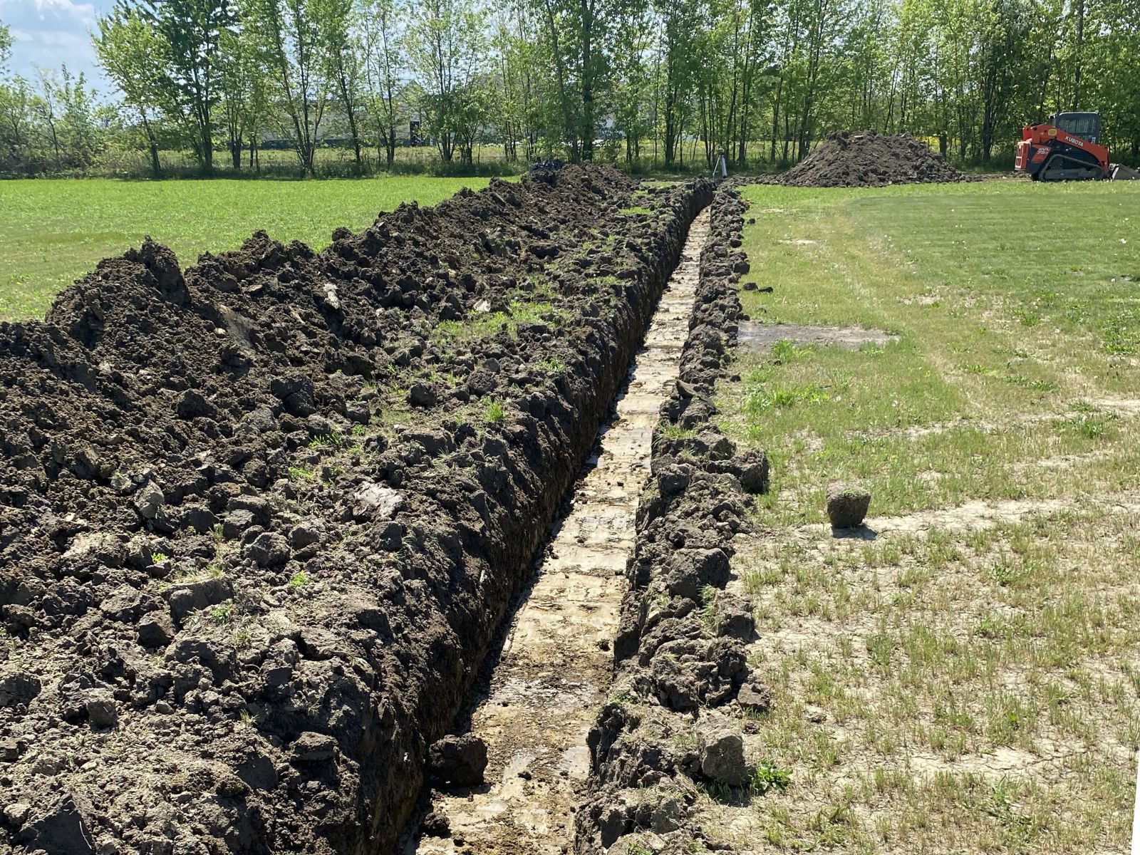 Long narrow trench dug in a grassy field with piles of soil alongside and a small orange excavator in the background.