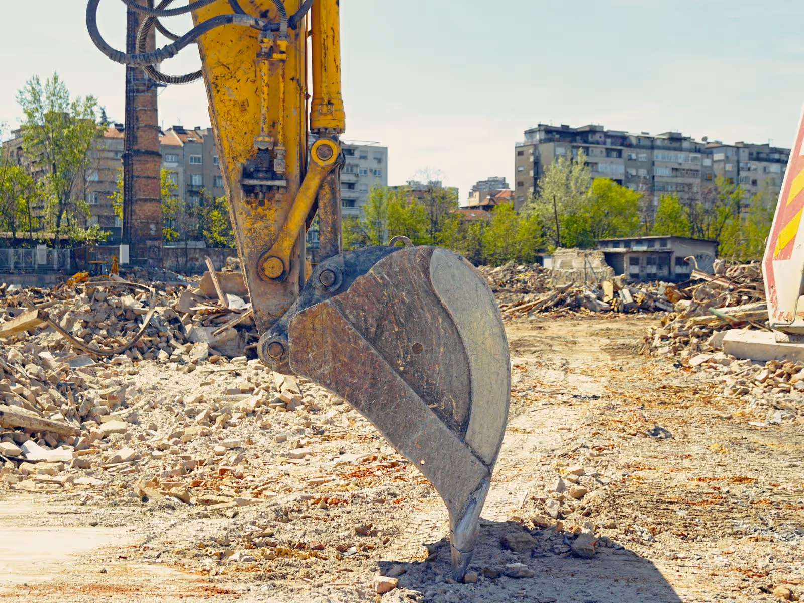 Close-up of a yellow excavator bucket at a demolition site with rubble and buildings in the background.